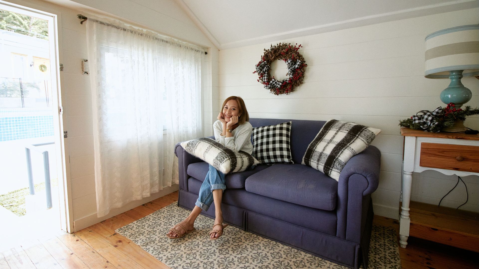 A cheerful woman sits comfortably on a sofa in a warmly decorated living room.
