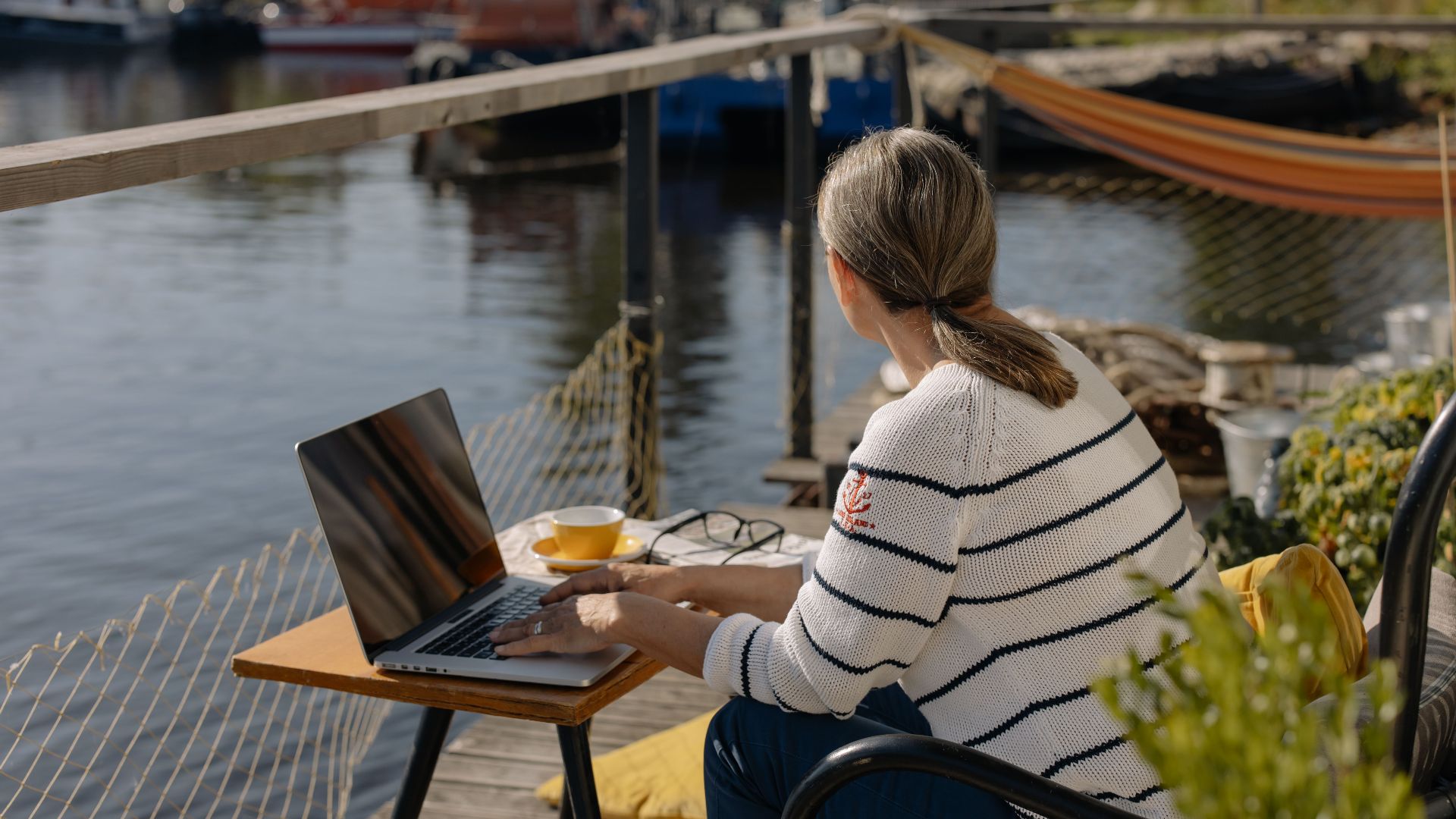 Woman working remotely on a laptop by the waterfront, enjoying a peaceful outdoor setting.