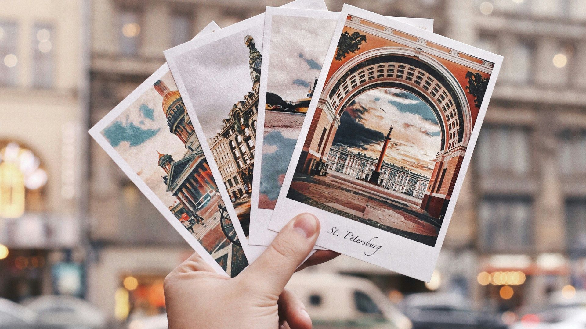 A hand holds postcards in front of a historic building in Saint Petersburg, Russia.