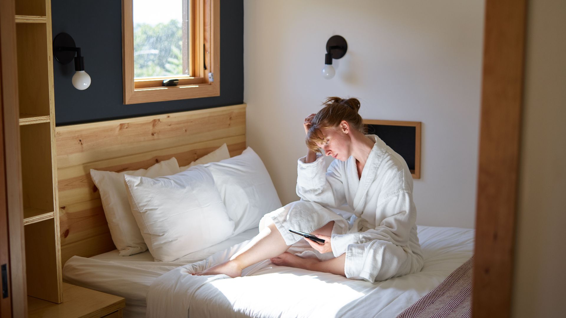 Full length barefoot focused female in white warm bathrobe chilling on cozy bed with ebook in modern bedroom on sunny day