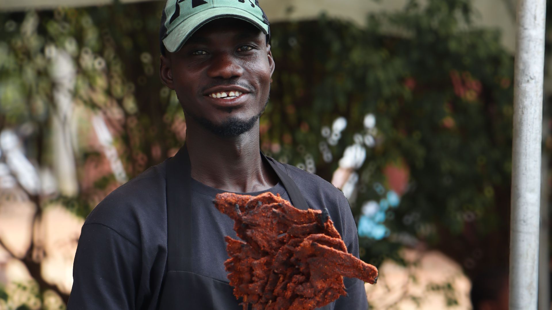 Man grilling traditional suya skewers outdoors at a food stall, smiling and serving customers.