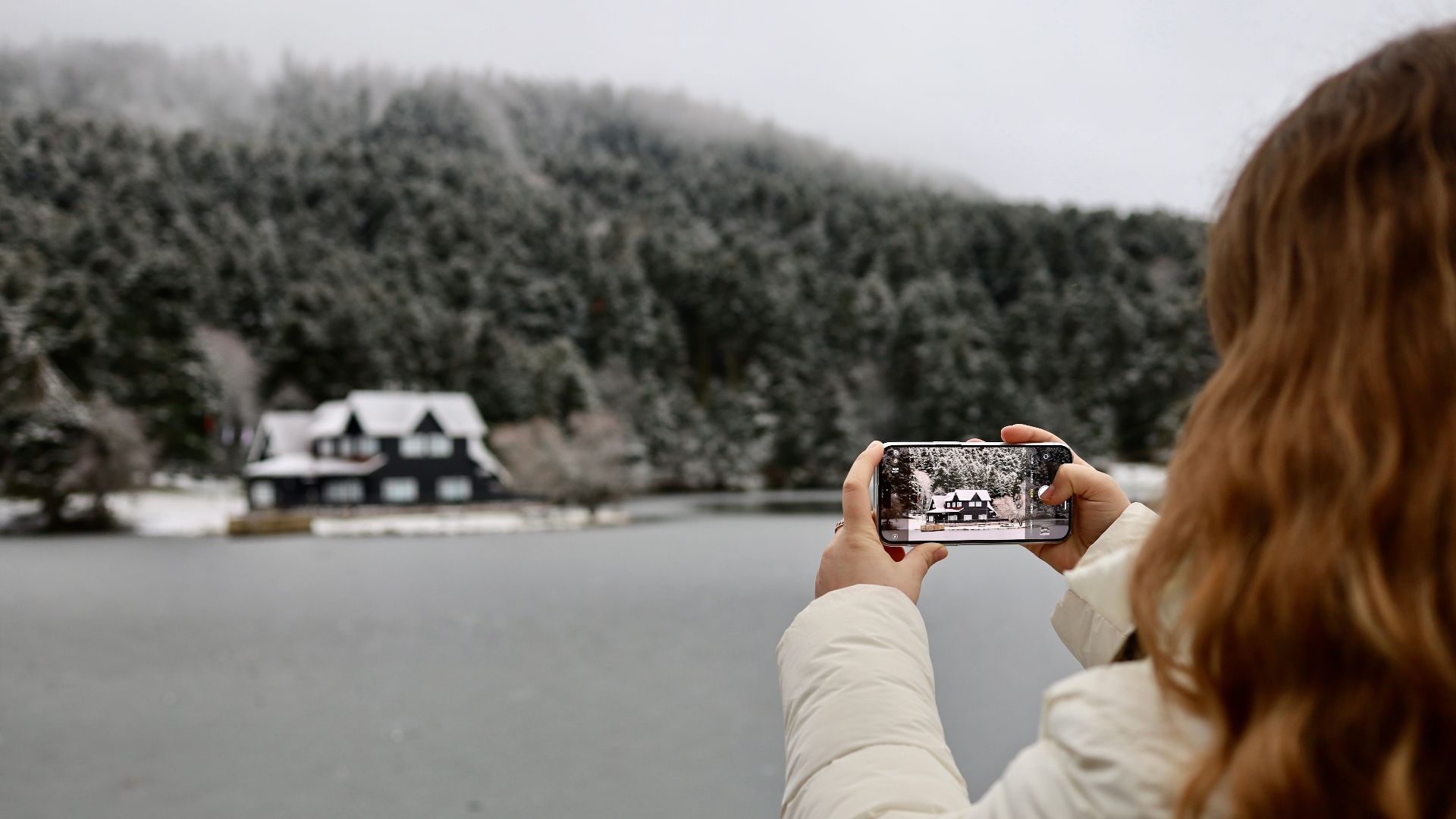 Woman photographing a snowy lake house near forested mountains in Bolu, Turkey.