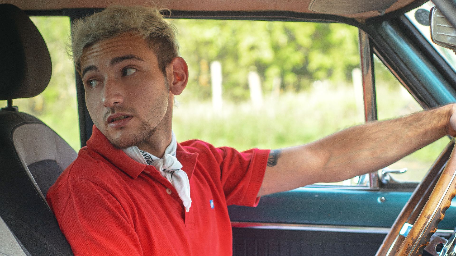 Young man in red polo shirt driving a classic car, looking attentively outside. Captured on a sunny day.