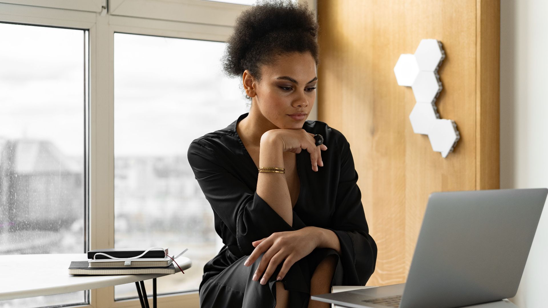 A woman in a black dress working remotely using a laptop by the window in a modern room.