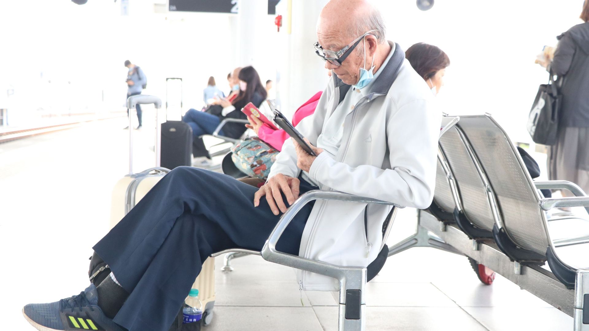 Senior adult using a smartphone in an airport terminal, sitting with luggage nearby.