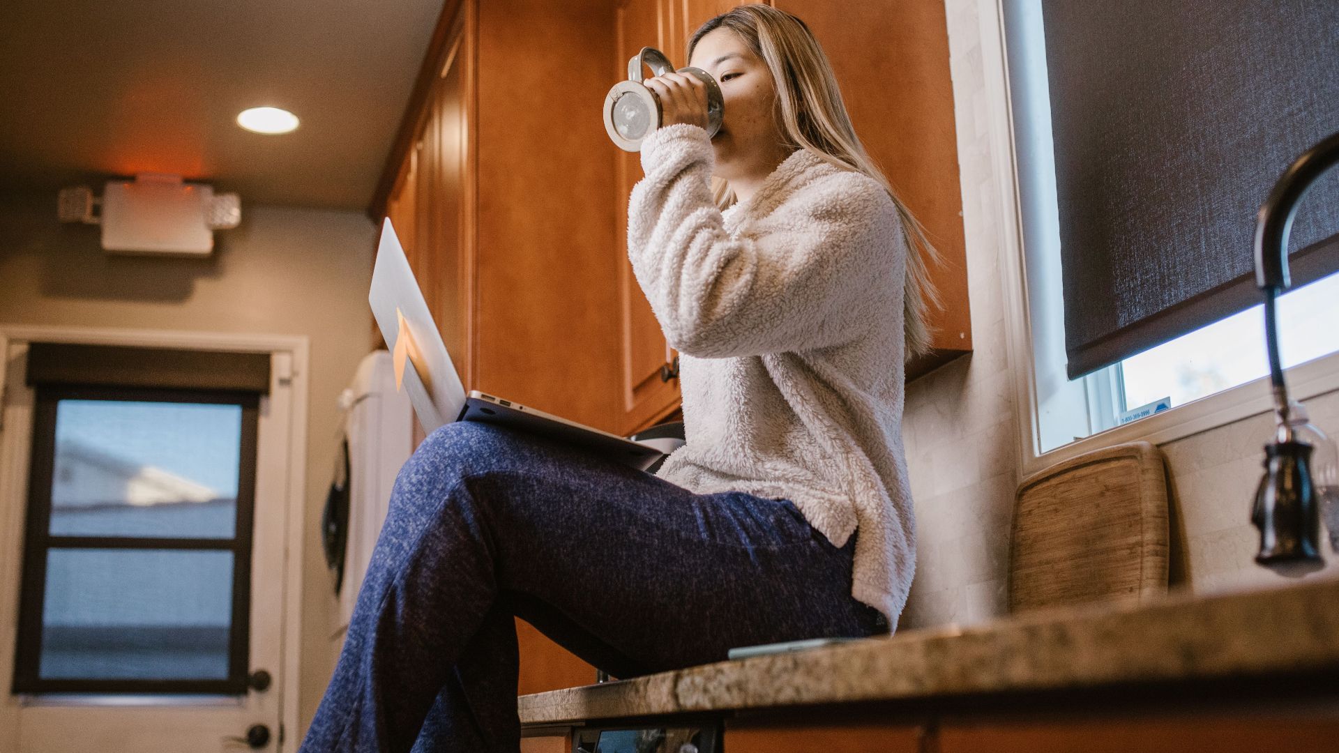 Casual morning scene of a woman enjoying coffee and using a laptop sitting on a kitchen counter.