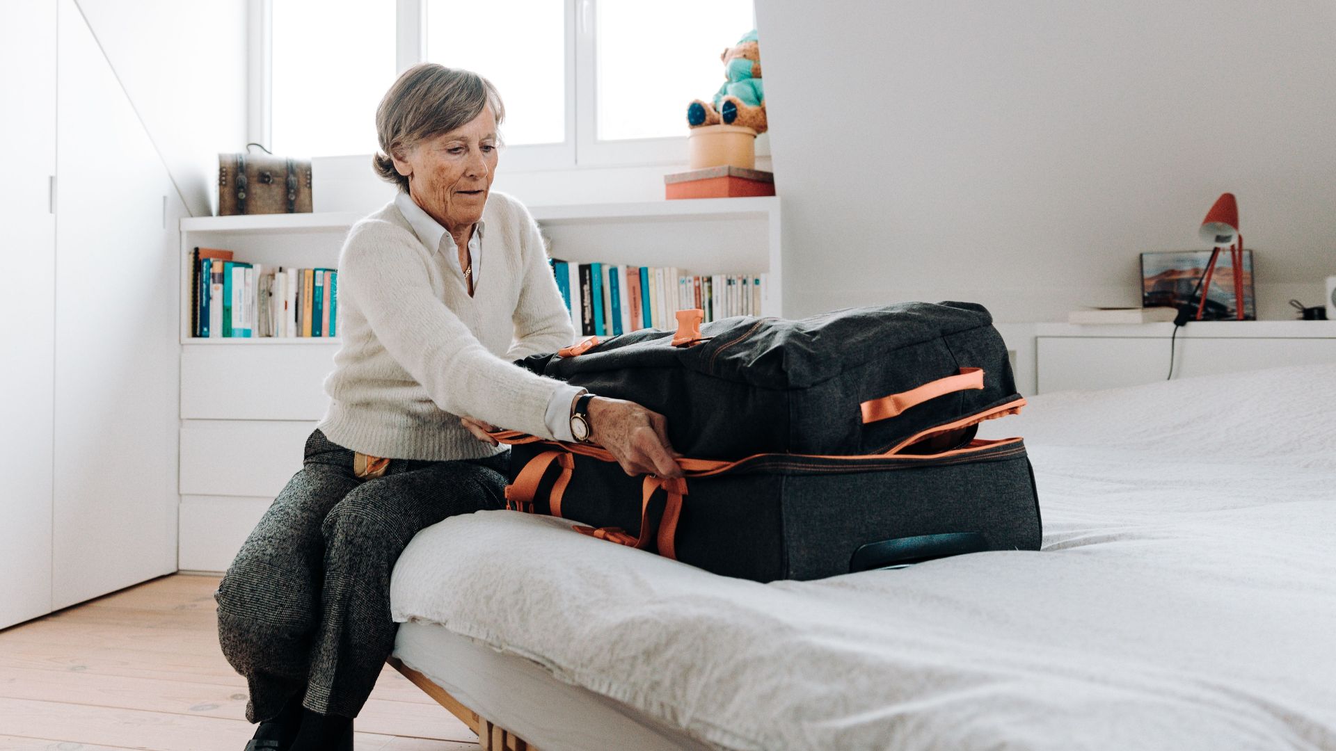 Elderly woman packing suitcase on bed in bright, modern bedroom setting.
