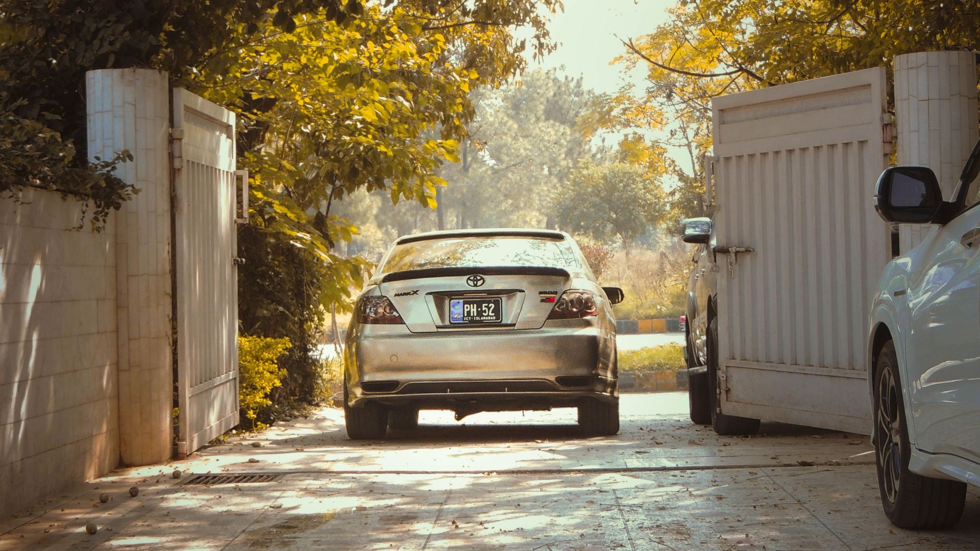 A sports car parked in a sunlit, leafy driveway during fall, framed by open gates.