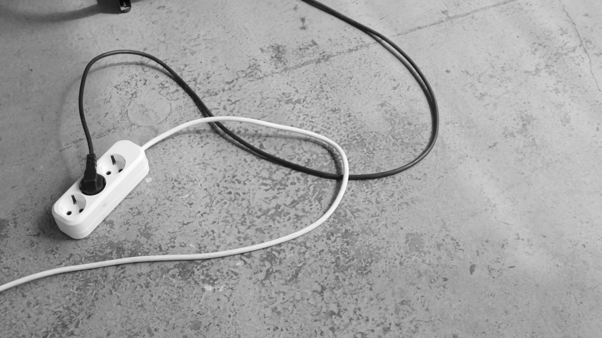 A minimalist black and white image of a floor fan and power strip on a concrete floor.