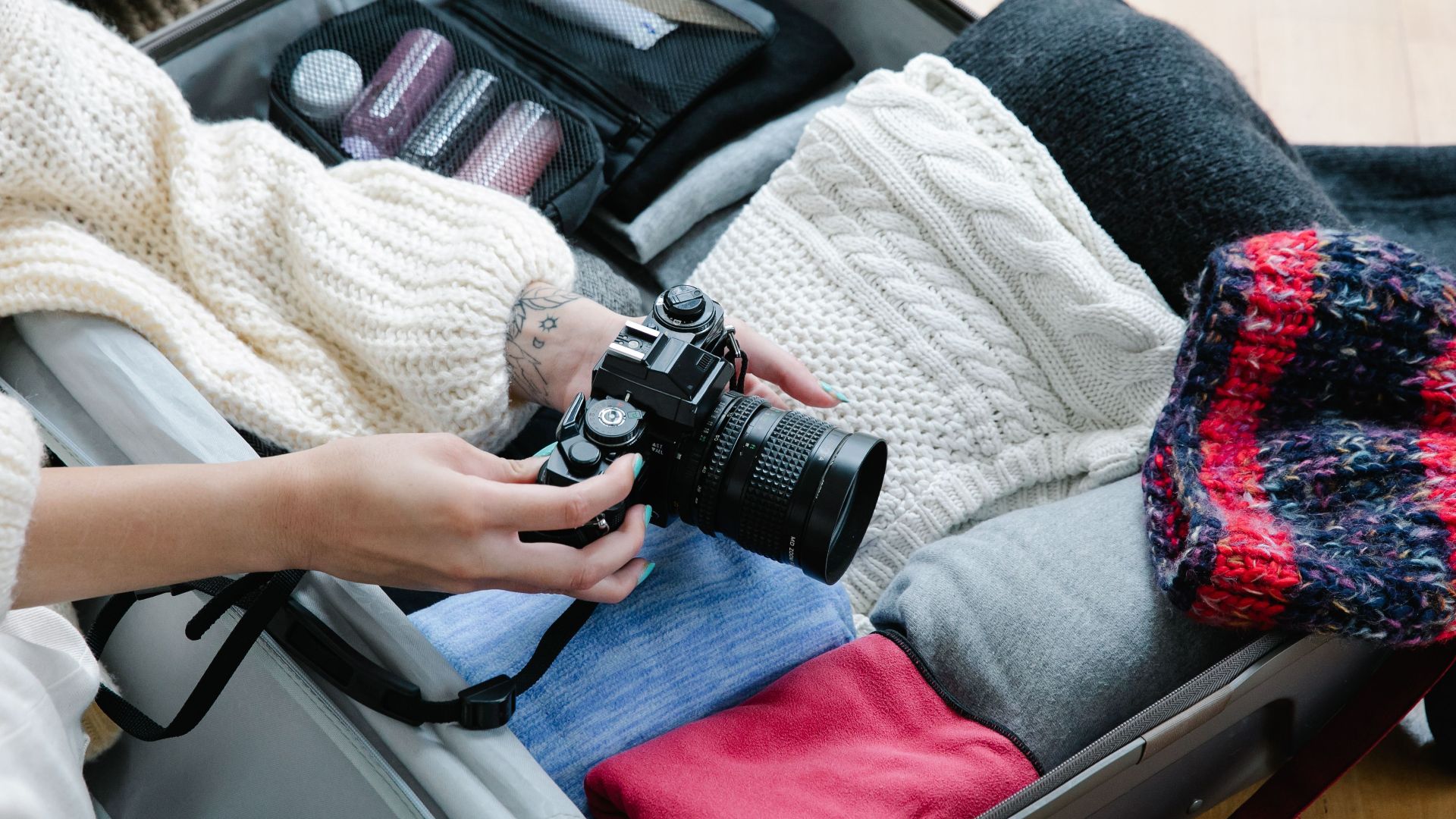 A person packing sweaters and a camera in a suitcase, preparing for travel.
