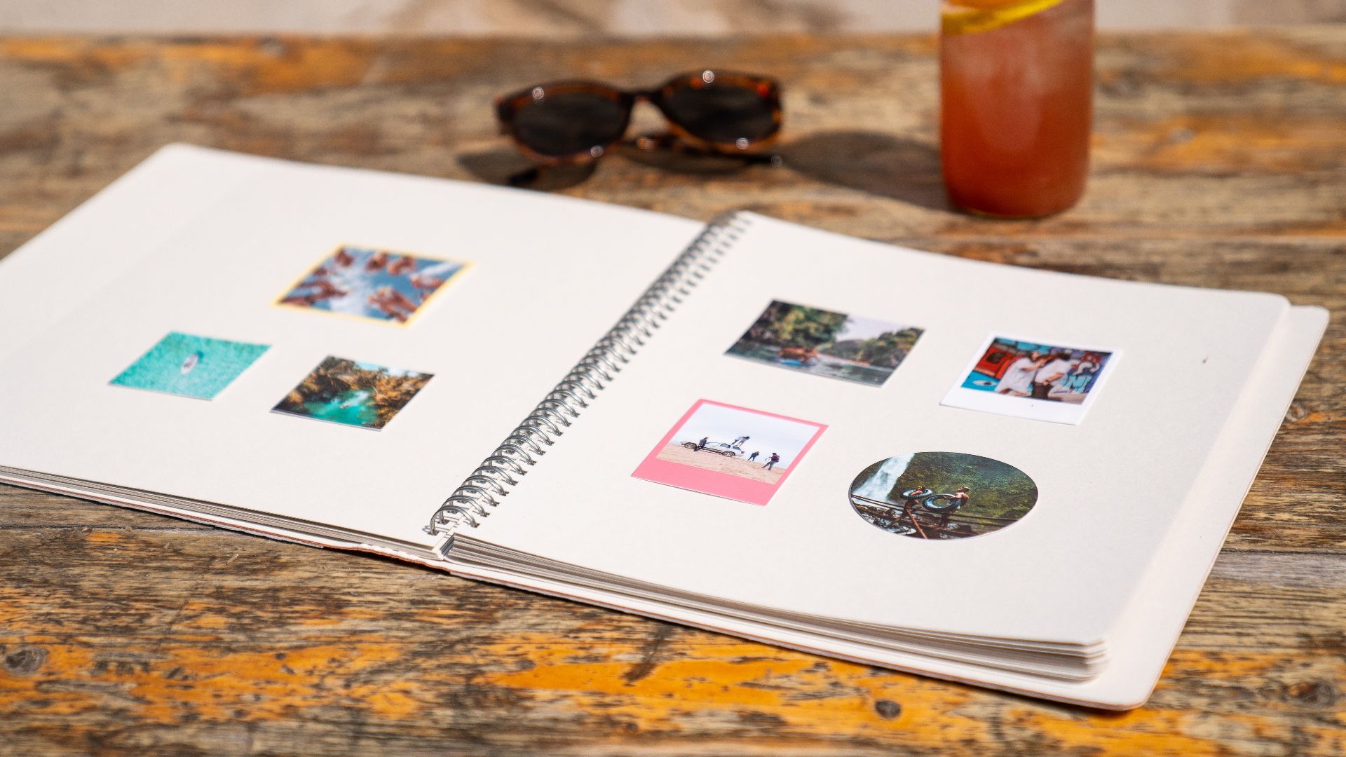 Open photo album on a wooden table with sunglasses and drink, evoking summer memories.