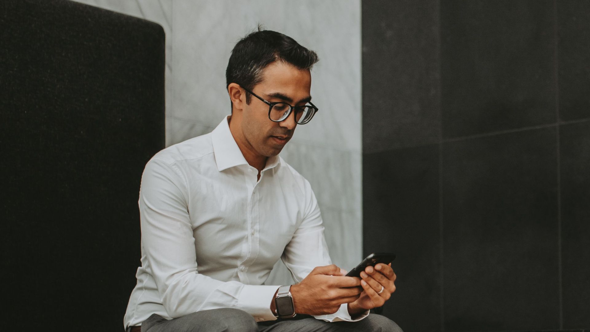 A businessman in a modern lounge checks his smartphone while wearing a white shirt and glasses.