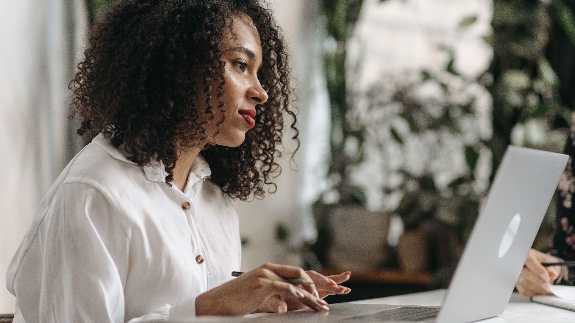 Young woman with curly hair typing on laptop in a bright, plant-filled office space.