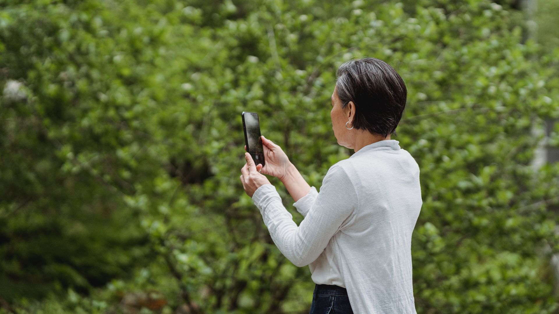 An adult woman captures a photo with her smartphone amidst a beautiful green landscape.