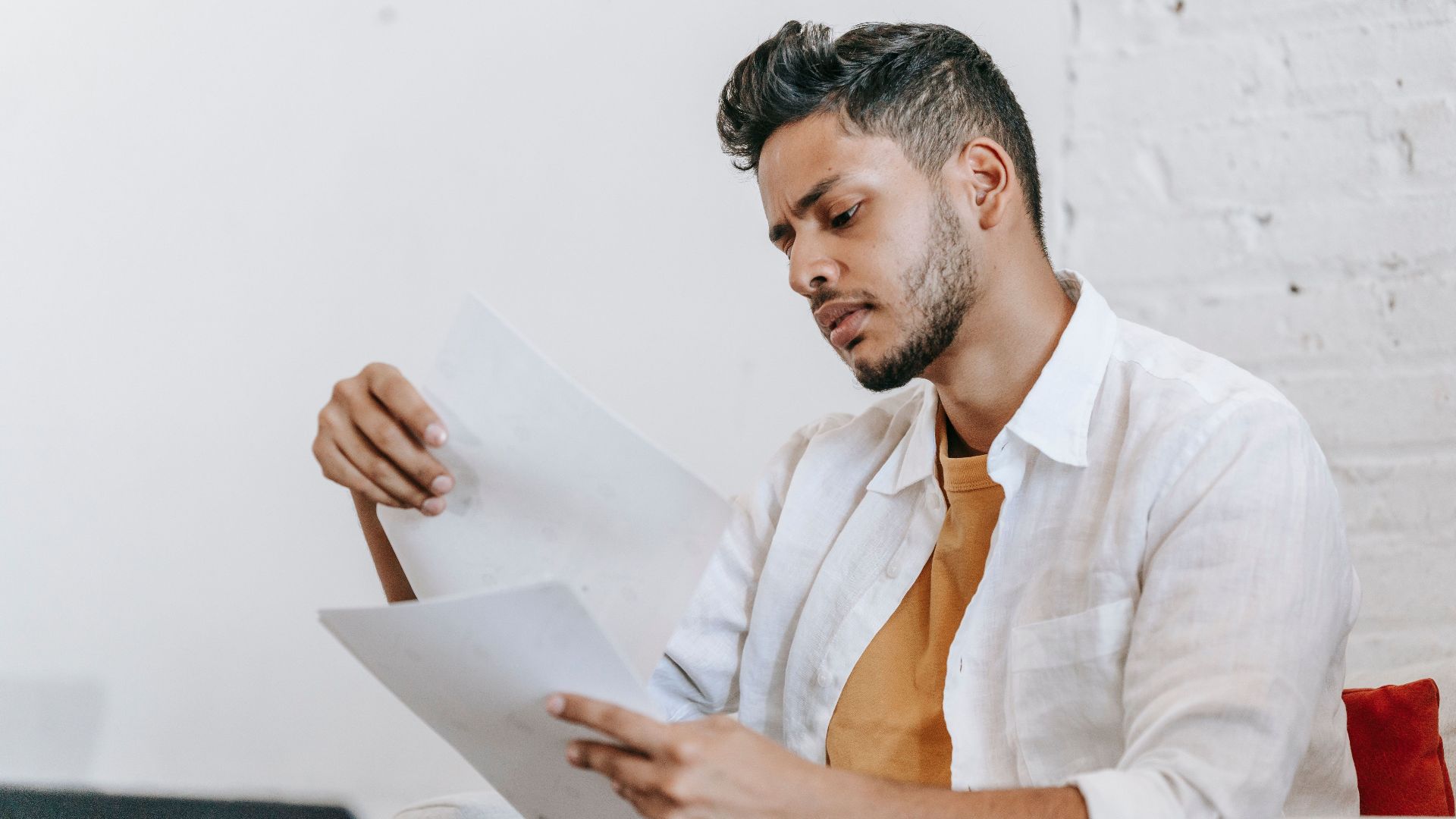 Concentrated ethnic male in casual wear reading documents and sitting on armchair near opened netbook