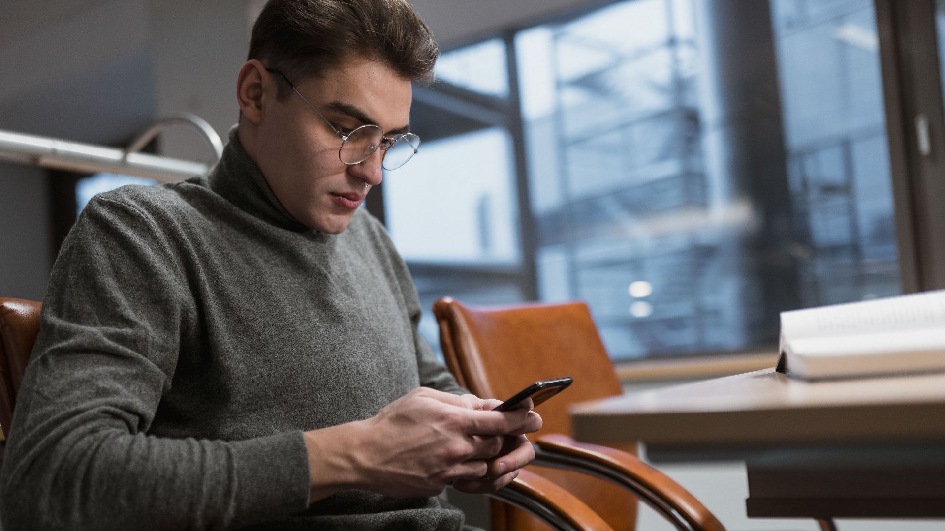 A young man in a grey sweater using a smartphone at an office desk.