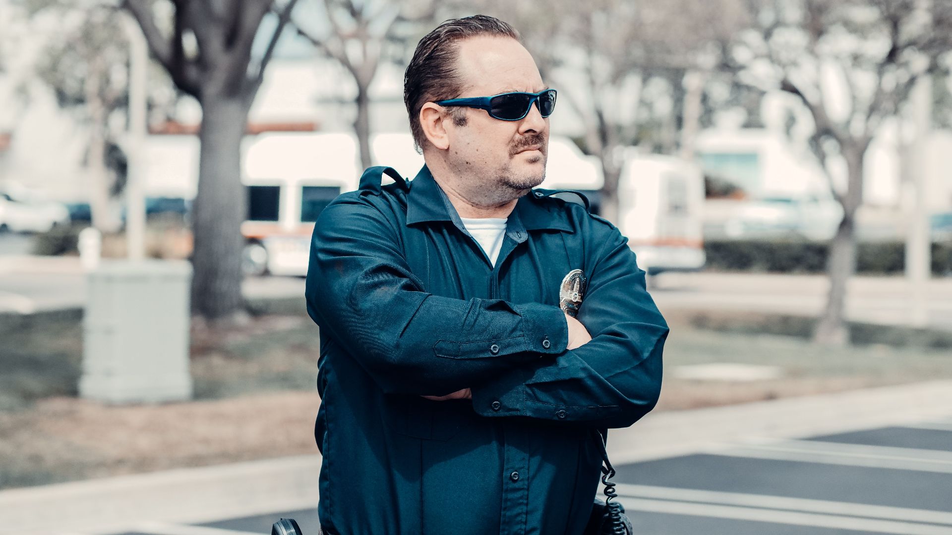 A police officer in uniform stands confidently outdoors with arms crossed and sunglasses.