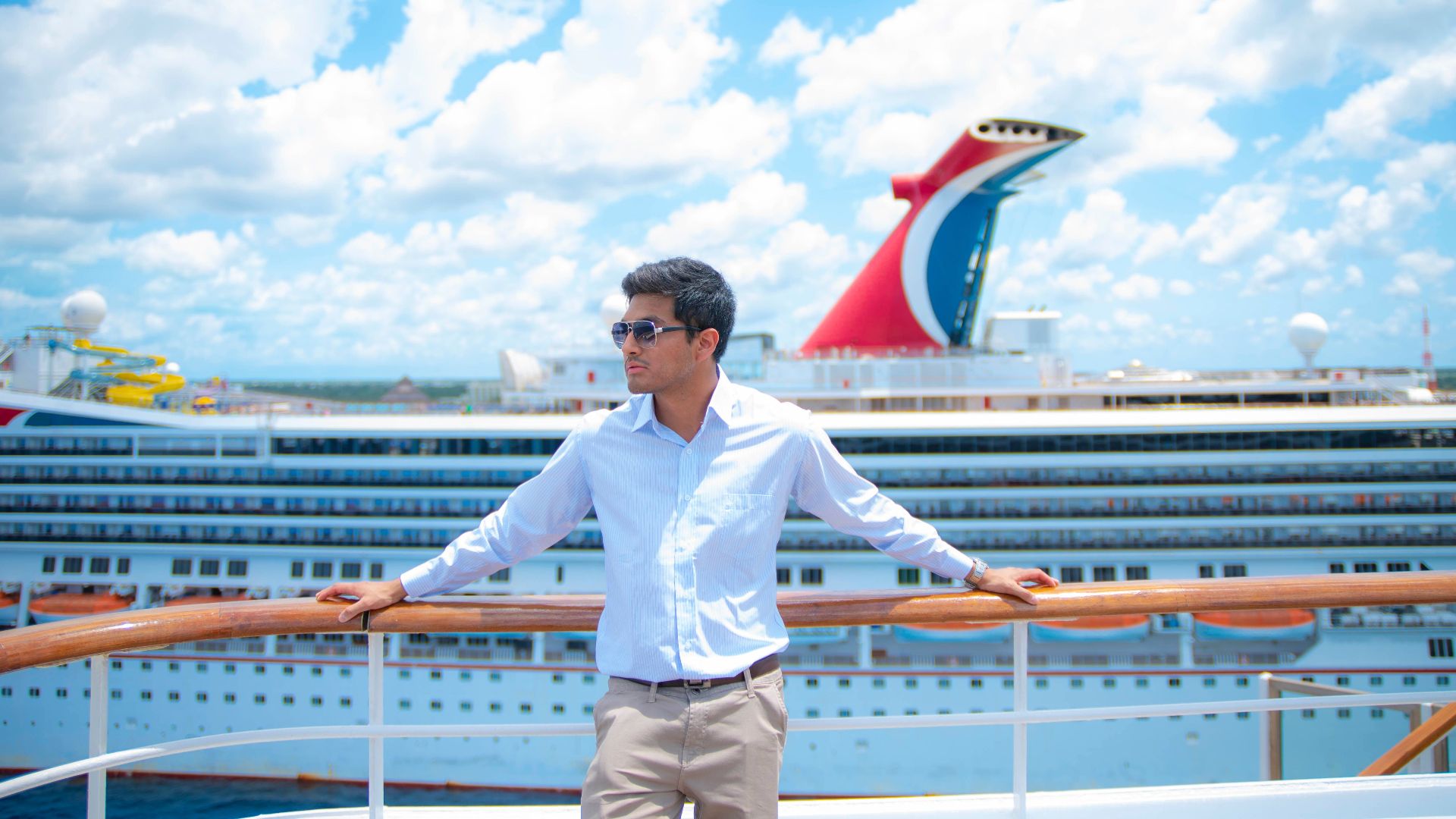 A man in sunglasses and white shirt enjoys a sunny day on a cruise ship deck with puffy clouds overhead.