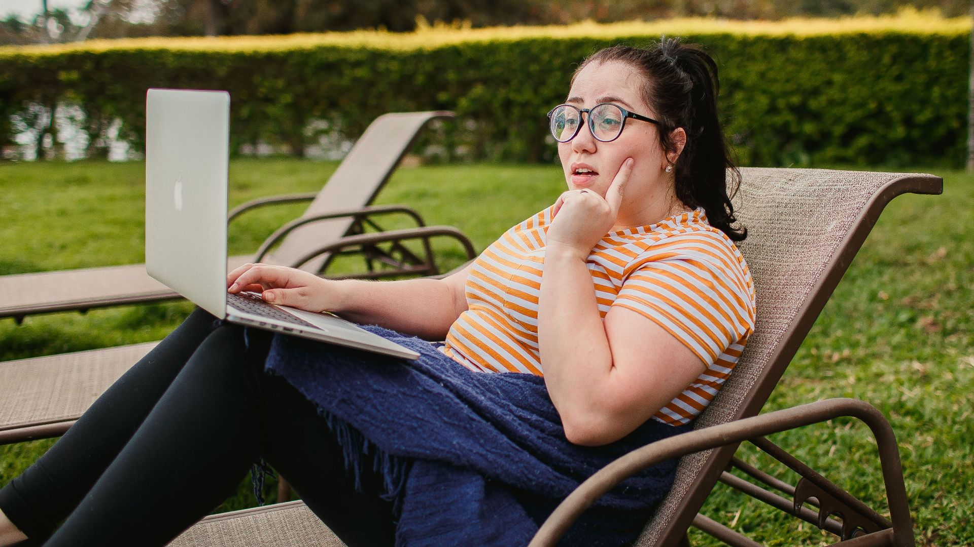 Woman relaxing on lounge chair with a laptop in a lush green garden, enjoying a sunny day.