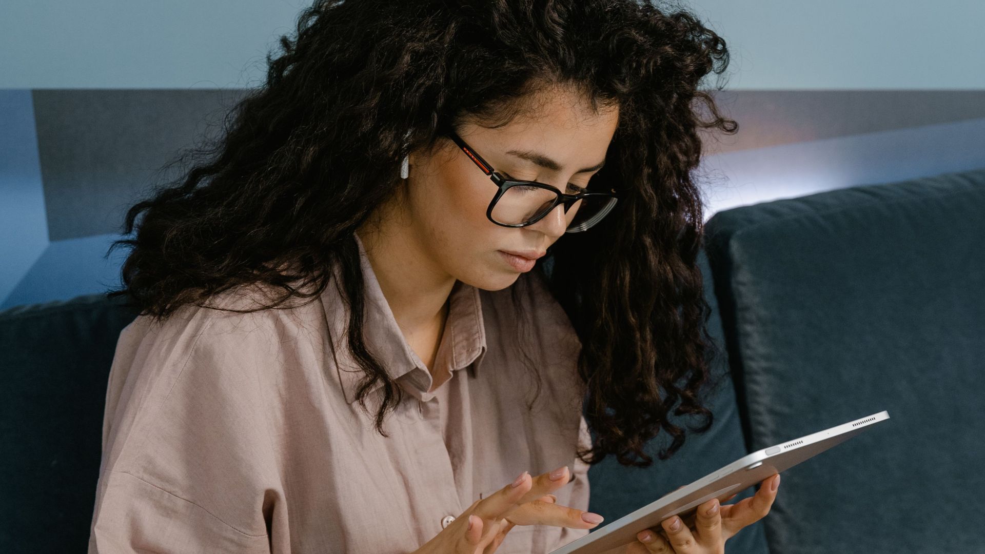 Young woman with curly hair and glasses using a tablet while sitting on a couch indoors.