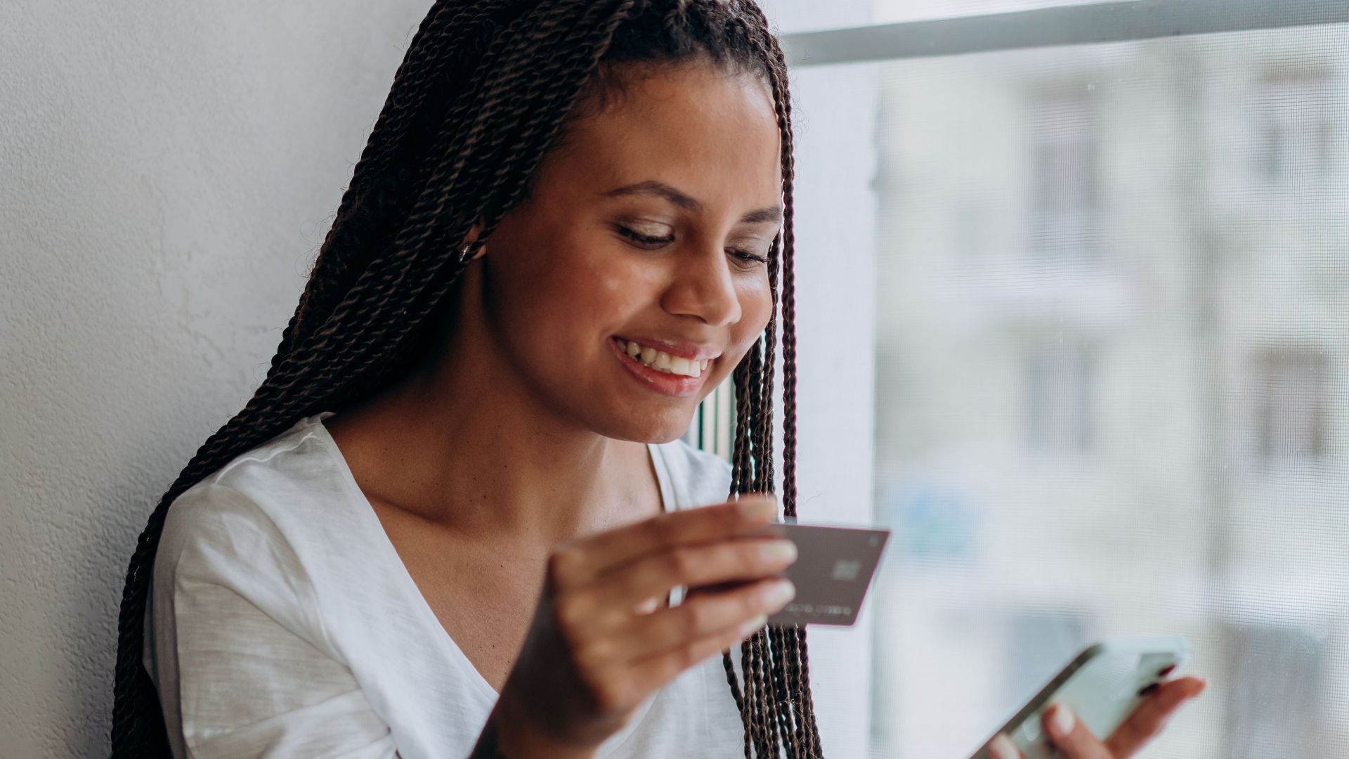 A smiling woman using a mobile phone and credit card for online shopping by the window.
