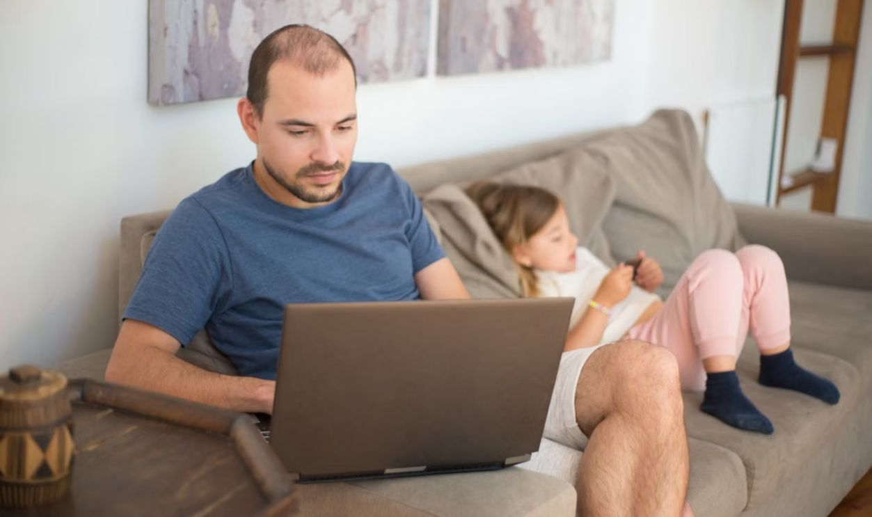 A man using a laptop while a child relaxes on the sofa. Cozy and modern living room setting.