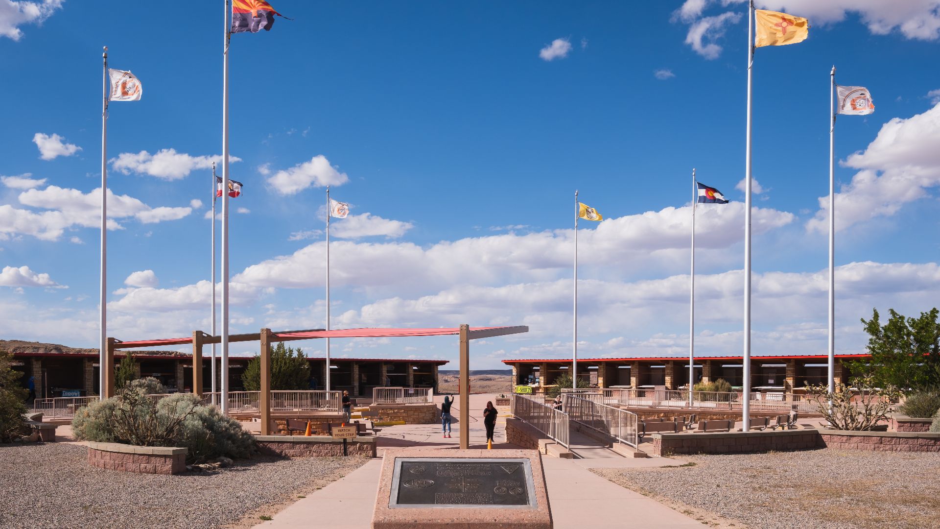 The concrete plinth bearing the bronze commemorative plaque on the access walkway of the Four Corners Monument, surrounded by the poles flying the flags of the four states and of the Native American nations.