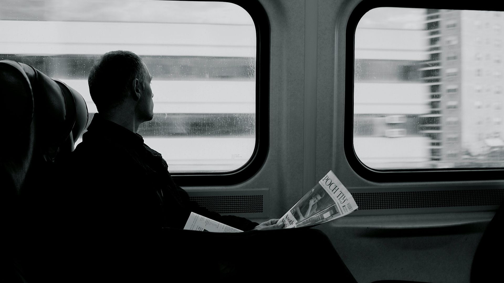 man inside train looking on window while holding newspaper grayscale photography