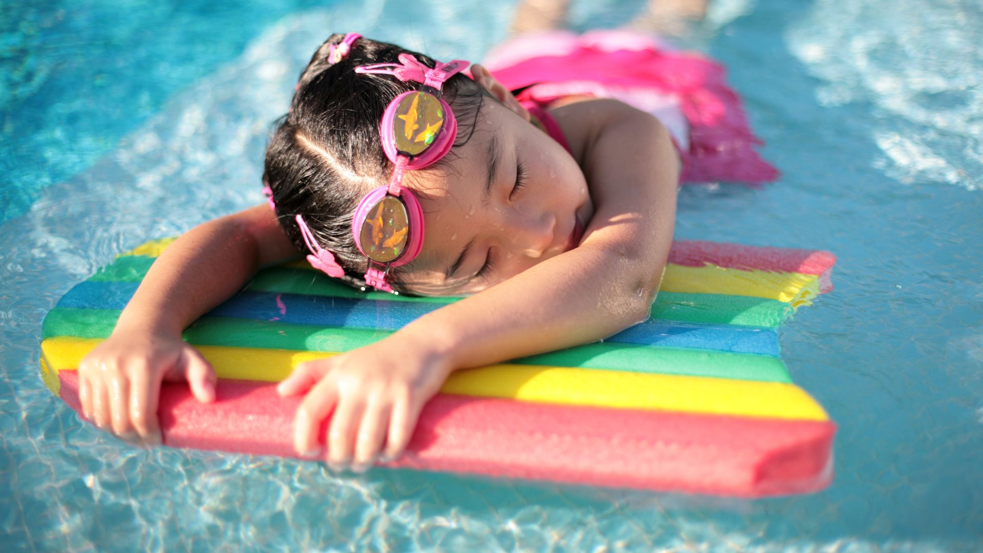 A Chinese girl taking a break in a swimming pool, grabbing on to a rainbow-coloured foam flotation device, probably polyethylene foam.