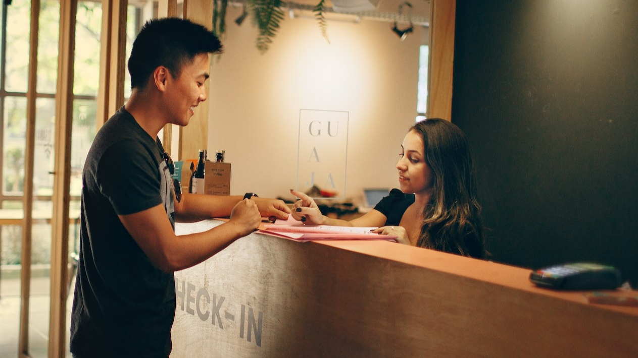 Man is standing in front of a front desk and talking with young woman with long hair.