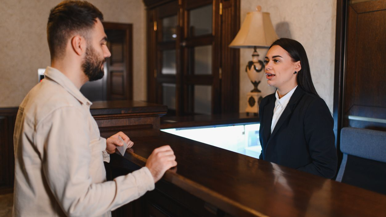 Shutterstock-2714036827, Guest standing at the hotel reception desk, talking to a female receptionist during a vacation or business trip