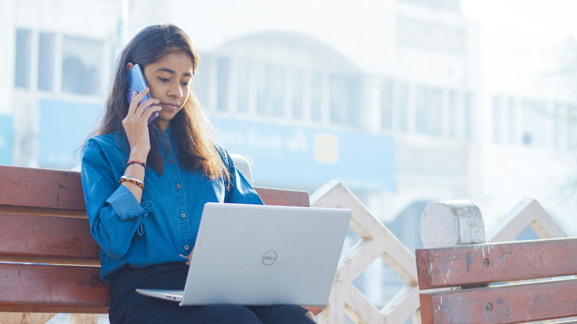 a woman sitting on a bench talking on a cell phone and using a laptop