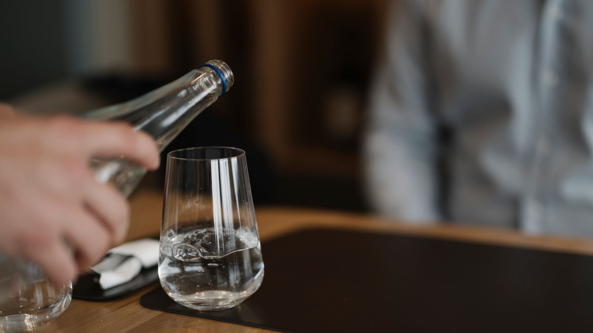 A waiter pours water from a bottle into a glass at a restaurant table. A person sits nearby, waiting for their drink. It is evening and the setting is warm and inviting.