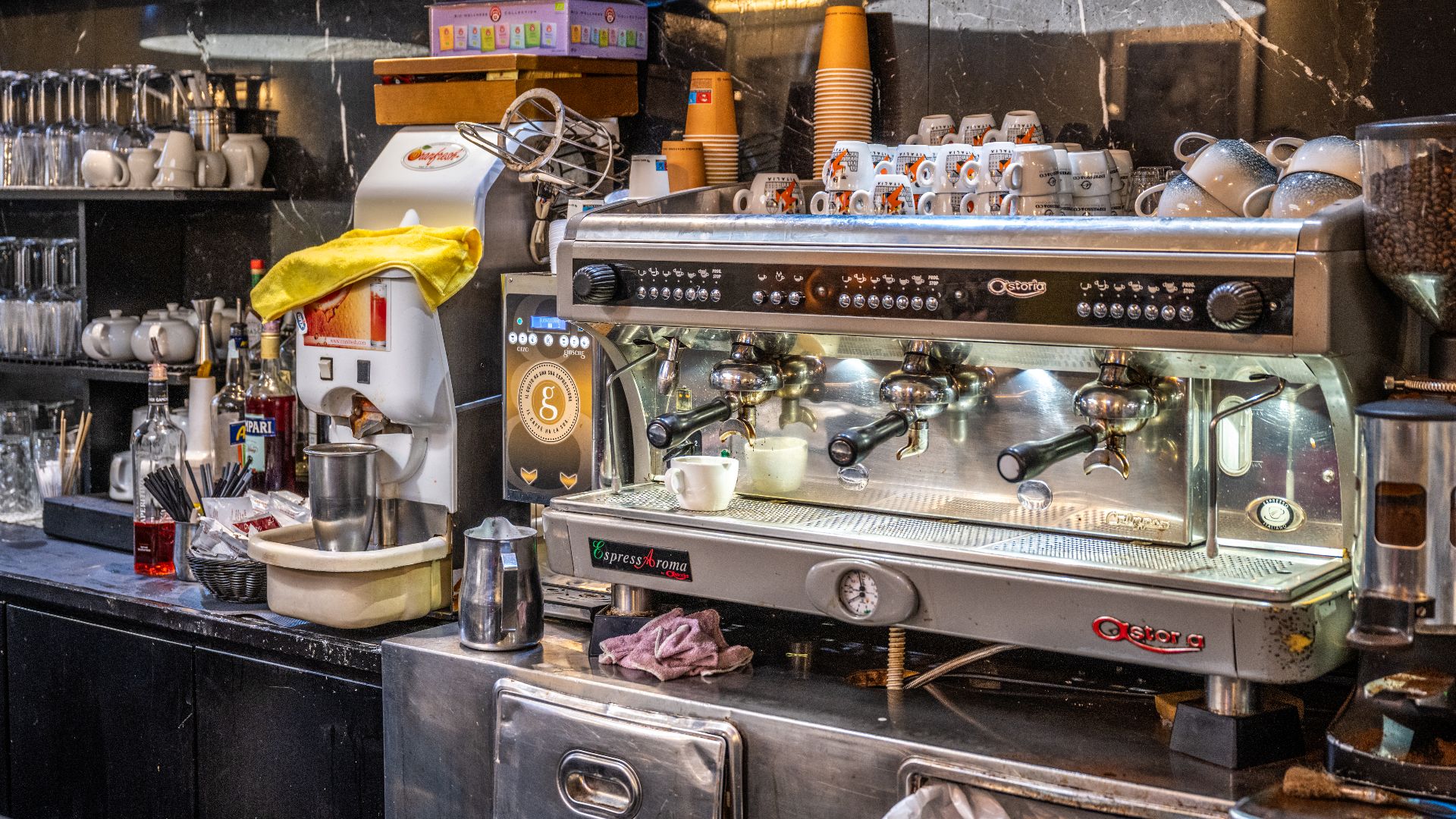 Counter of a coffee bar in Rome with a three group espresso machine, cups stacked on top to warm, a grinder, a juicer and bottles behind.