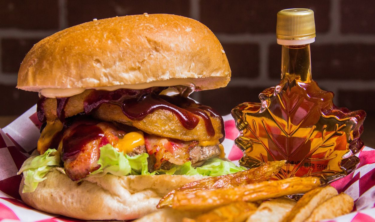 A mouthwatering burger stacked high with juicy meat, cheese, lettuce, and topped with barbecue sauce served alongside a bottle of maple syrup, showcasing comfort food at its best.