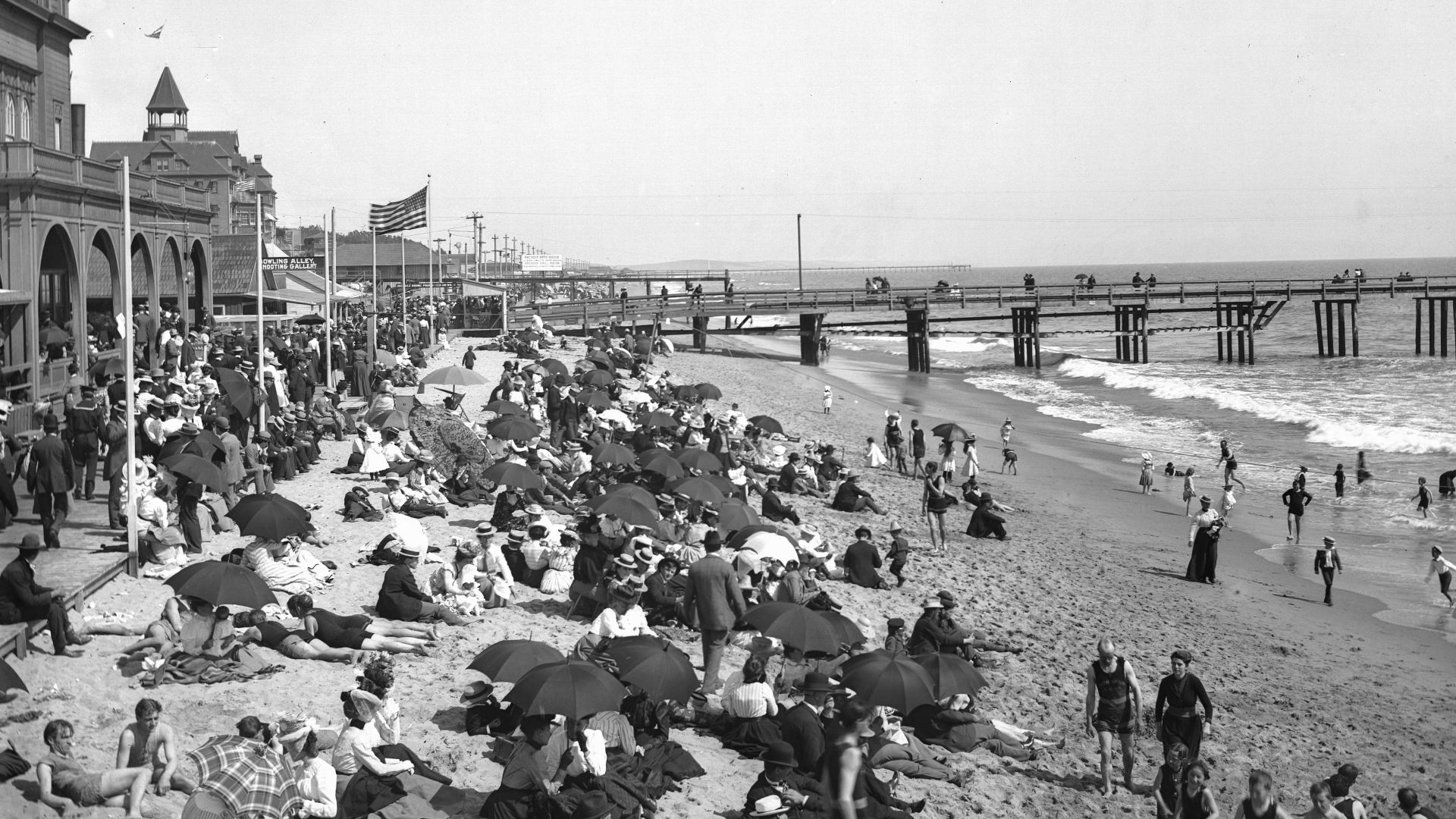 Crowds of people on Santa Monica Beach, ca.1900Photograph of crowds of people (mostly bathers, many with umbrellas) on the Santa Monica beach, ca.1900. A row of buildings lines the boardwalk. A pier is visible behind the people. An American flag flies fr