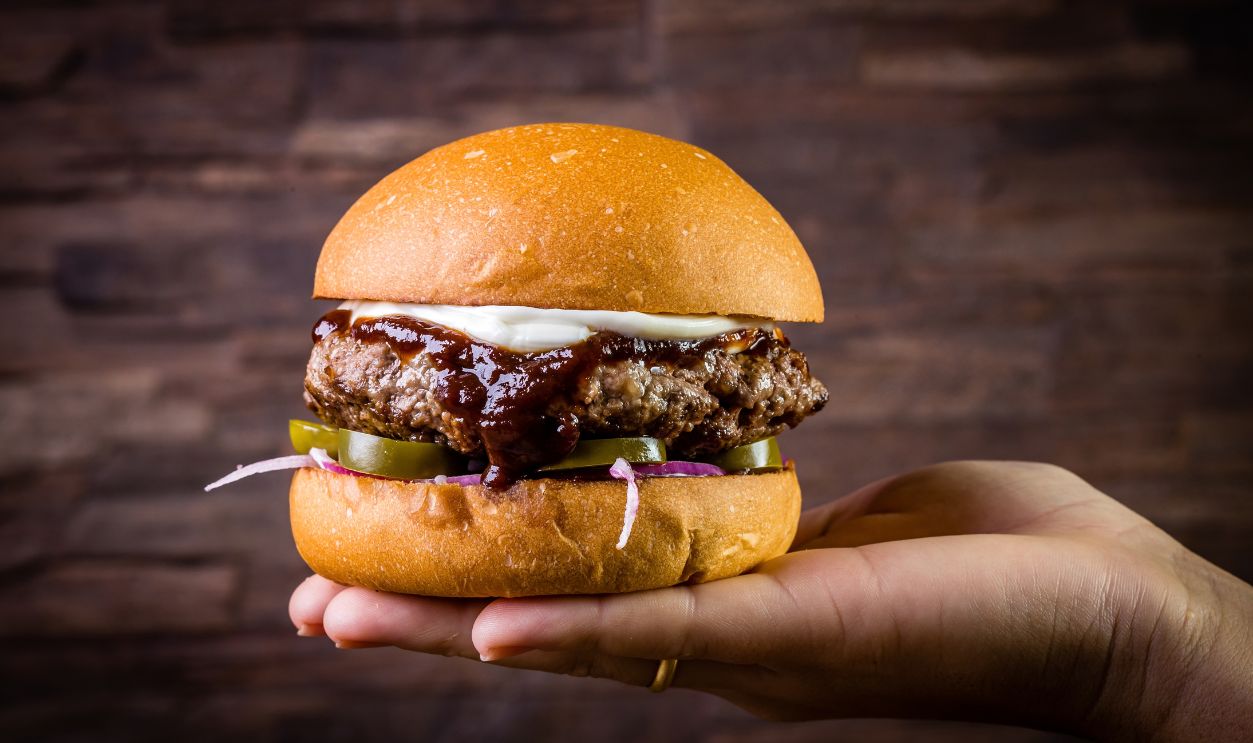 Hand holding a craft beef burger with cream cheese, purple onion, jalapeno pepper on wood table and rustic background