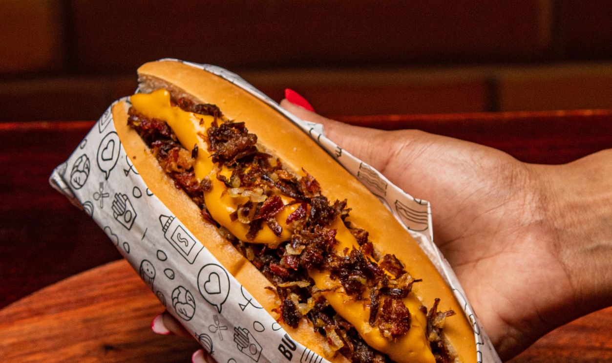 A close-up of a hand holding a gourmet hot dog with toppings on a wooden table