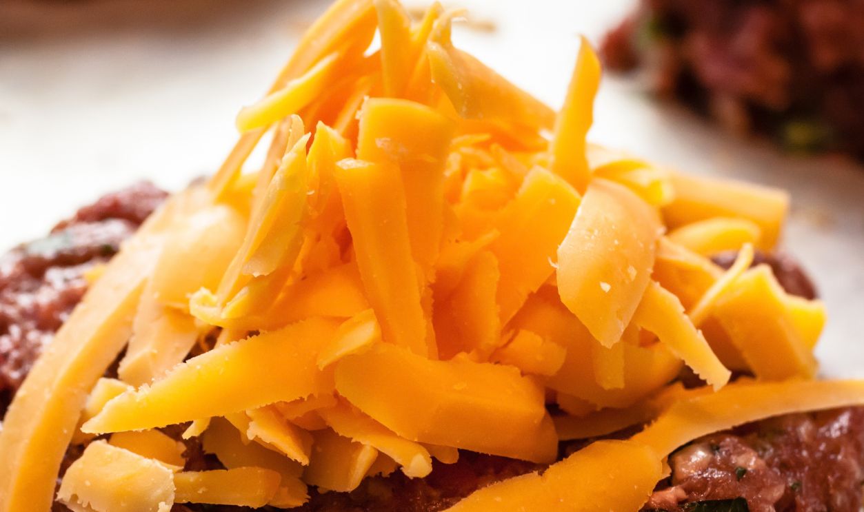 Close up of shredded cheddar cheese sitting on raw gourmet hamburgers, on silver plan waiting to be cooked