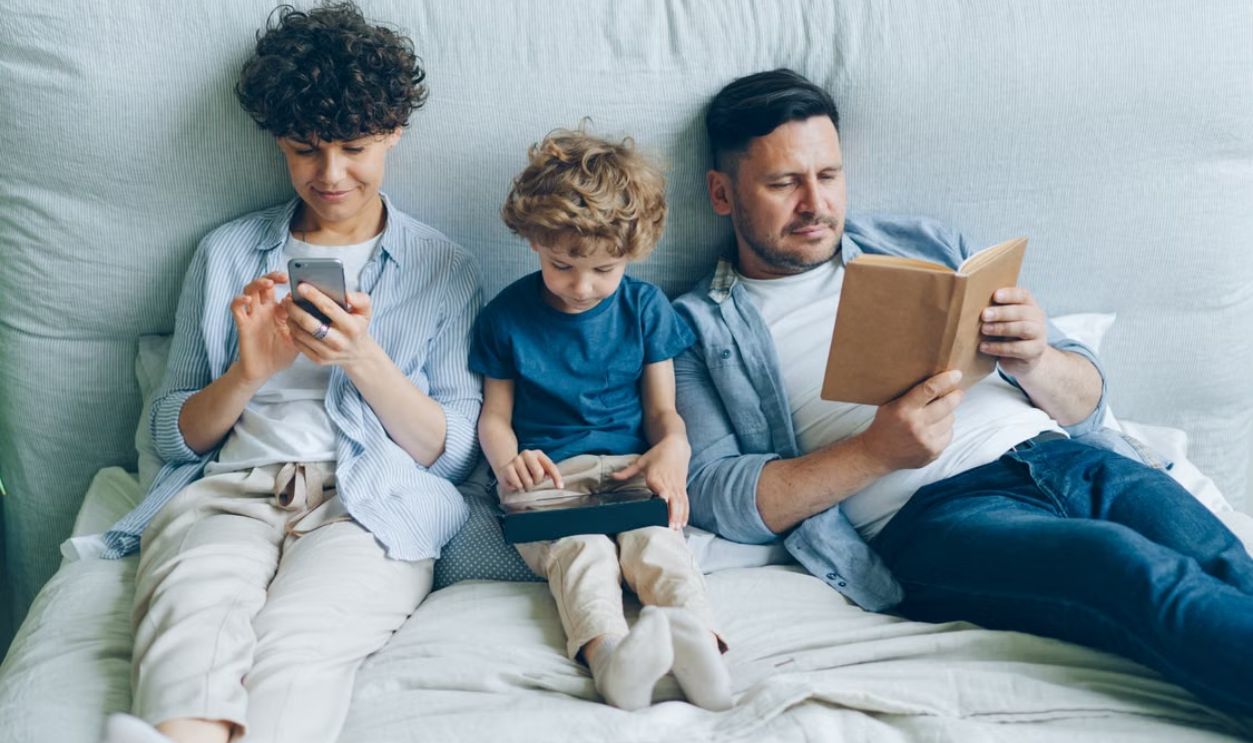A family relaxing on bed with a smartphone, tablet, and book, symbolizing modern family life.