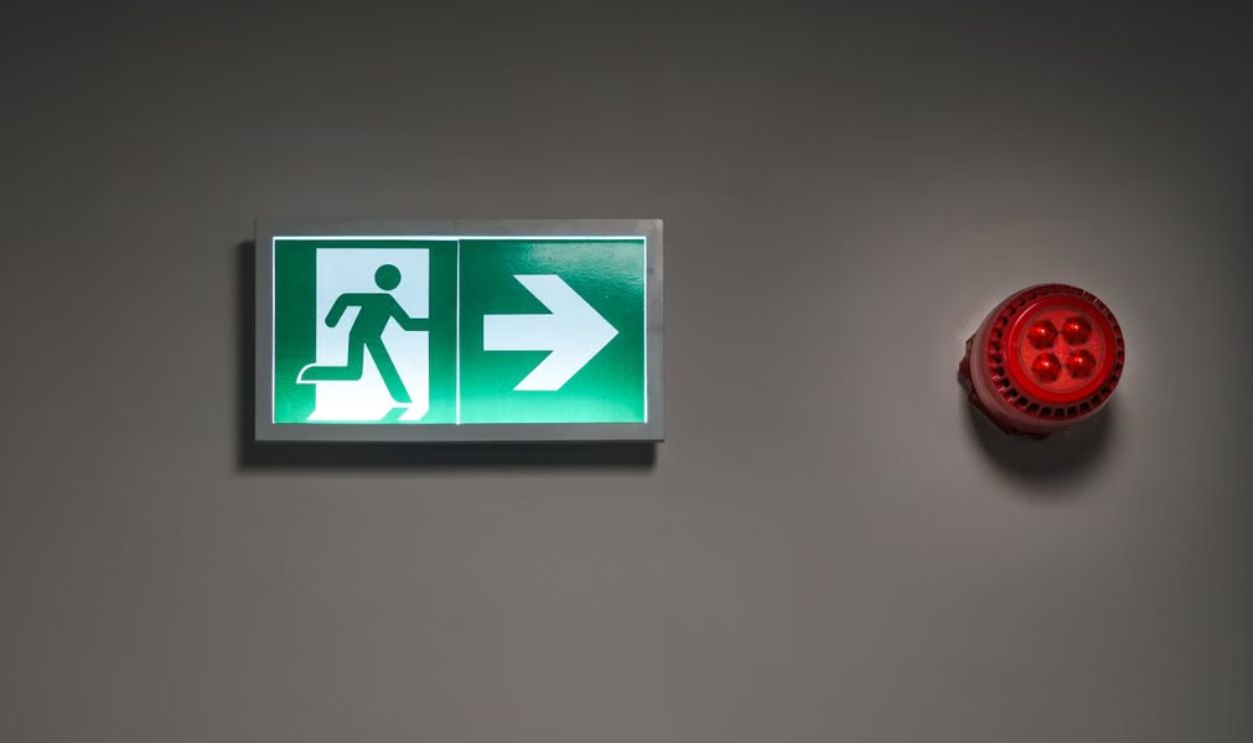 A brightly lit exit sign and alarm in an indoor corridor, indicating emergency escape route.