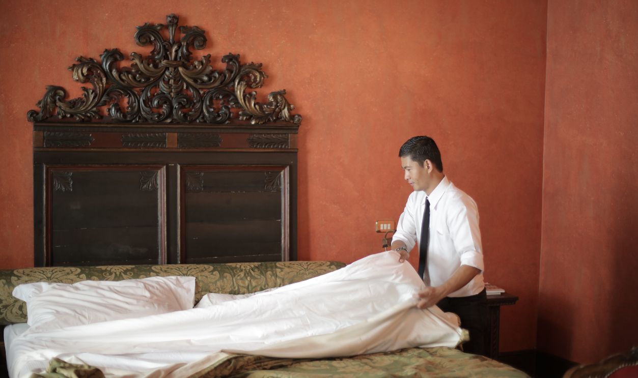 A hotel staff member in formal attire making a bed in a luxurious bedroom setting.
