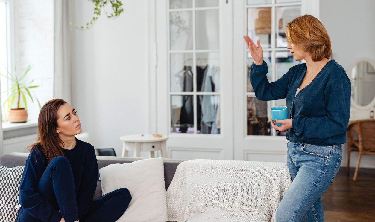 Two women engage in a friendly chat in a stylish living room, one sitting and one standing with a mug.