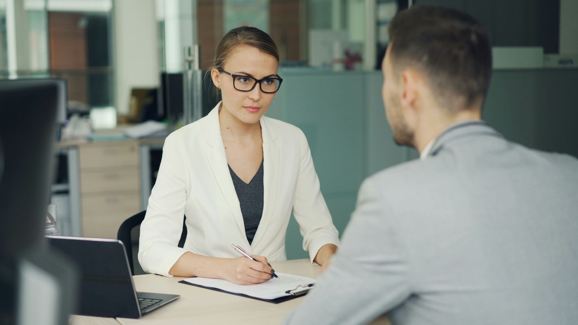 Woman in glasses interviews man at office desk.