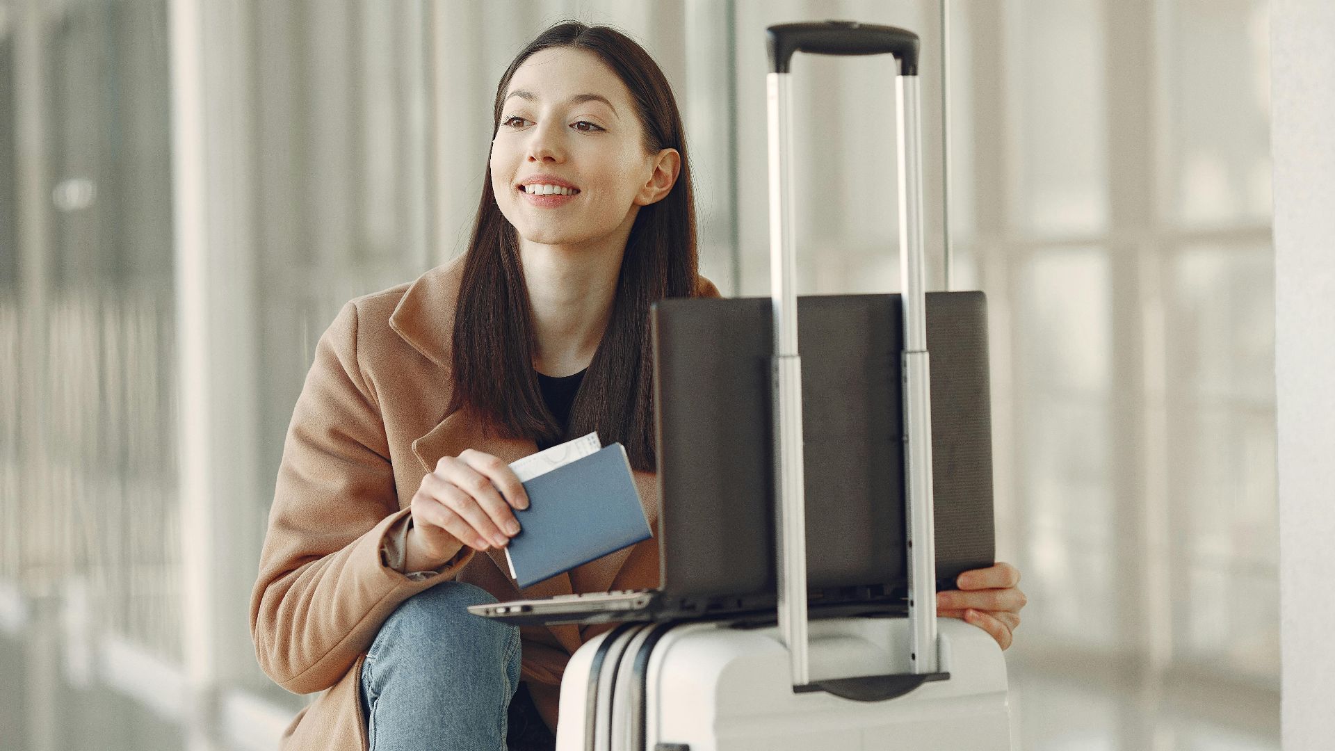 A woman sitting with her laptop and luggage, holding a passport, waiting in an airport terminal.