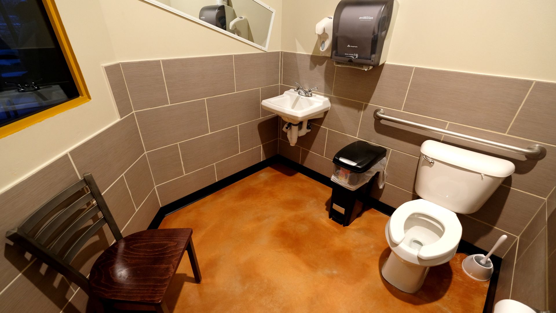 Chic City Bites restaurant bathroom that includes (clockwise, from left), a chair to place belongings, a two-way mirror to see what's going on in the dining room, a standard mirror, a sink, a liquid soap dispenser, a hands-free paper towel dispenser, a foot-operated trash can, a safety handrail, a toilet, a toilet brush, and a toilet paper roll.