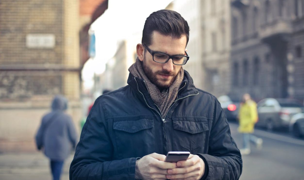 Man Wearing Black Zip Jacket Holding Smartphone Surrounded by Grey Concrete Buildings