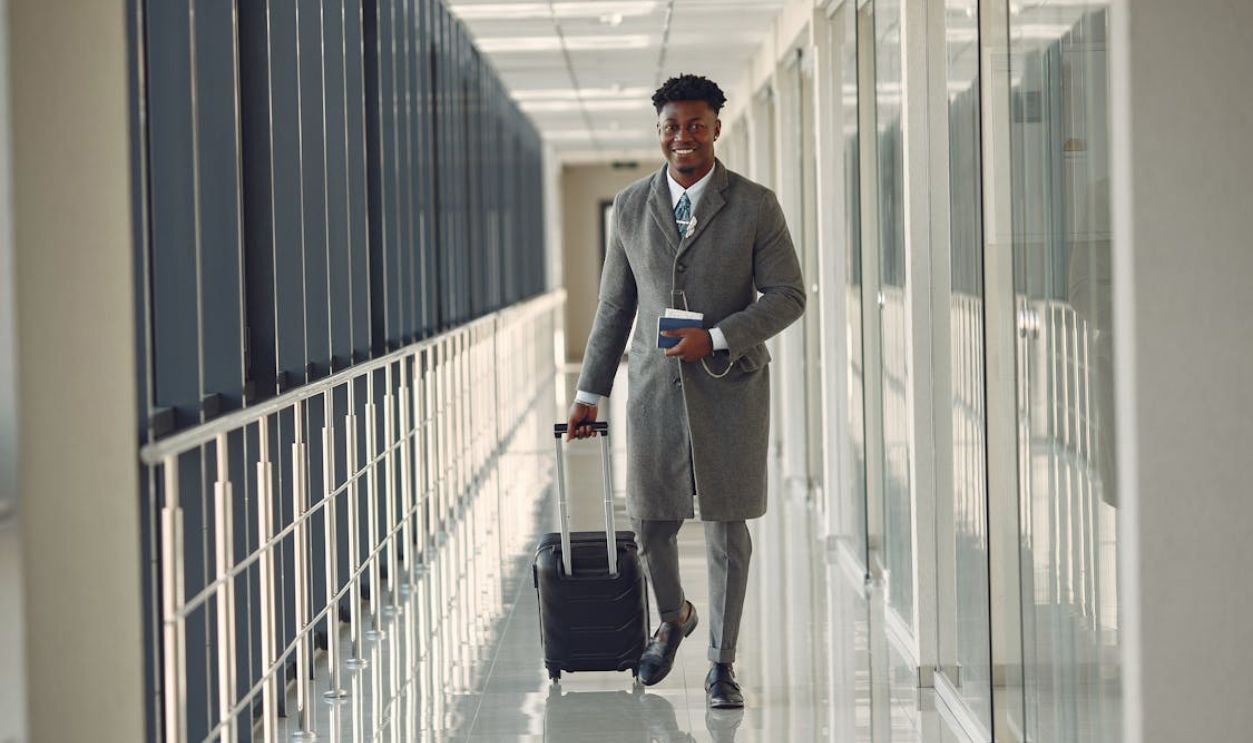 Stylish man with suitcase and passport walking along airport corridor