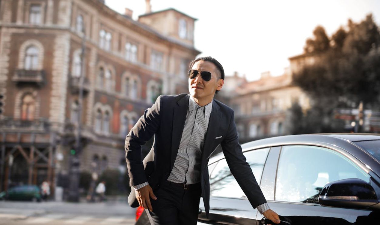 Photo of Man in Black Suit and Sunglasses Standing Beside Parked Car
