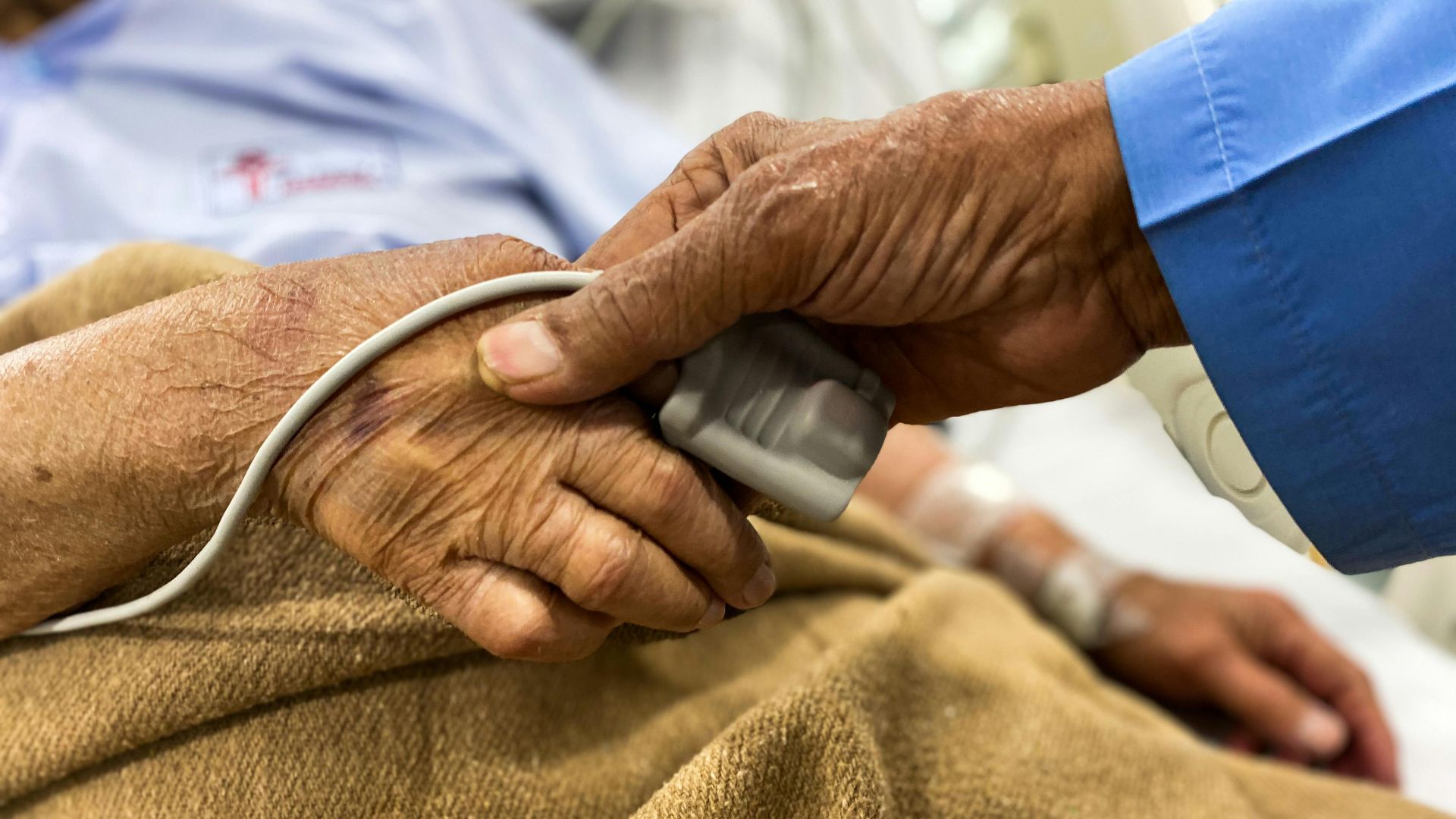 Crop old female patient lying on medical bed with pulse oximeter on finger and old man holding wrinkled hand